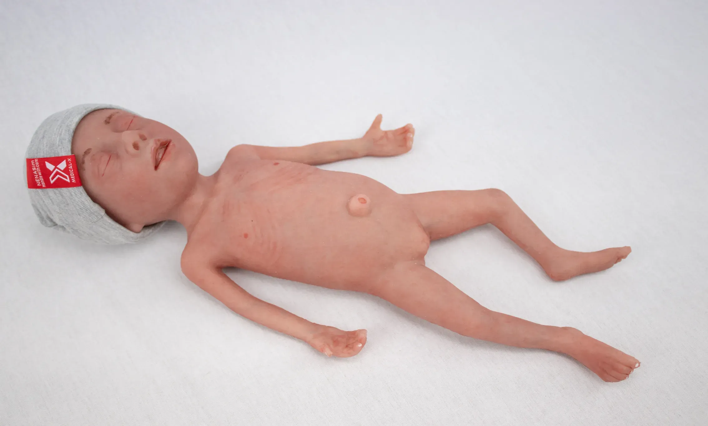 Simulador de bebé prematuro con gorrito gris colocado sobre superficie blanca.
